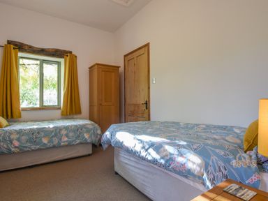 A bedroom with two single beds a wooden wardrobe a wooden door and a window with yellow curtains at Bradbridge Barn in Newton Ferrers