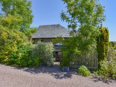 A garden with trees and bushes surrounding a wooden fence and stone house at Parsonage Farm Cottage in Newton Ferrers