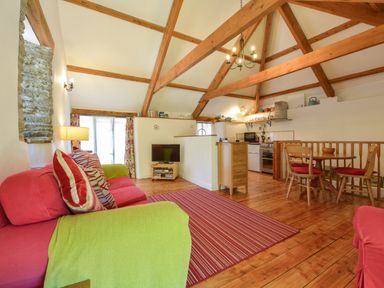 A living room with wooden beams a red sofa a small dining table and a kitchen at Parsonage Farm Cottage in Newton Ferrers