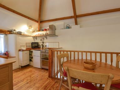 A kitchen area with wooden floors and ceiling beams containing a stove, oven, refrigerator, shelves with dishes and teapots, and a wooden dining table with chairs at Parsonage Farm Cottage in Newton Ferrers