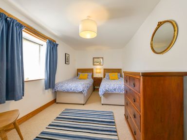 A bedroom with two single beds a wooden chest of drawers a striped rug and blue curtains at Parsonage Farm Cottage in Newton Ferrers