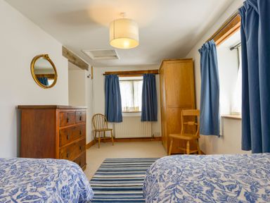 A bedroom with two beds, a wooden chest of drawers, two wooden chairs, a wooden wardrobe, blue curtains, and a striped rug at Parsonage Farm Cottage in Newton Ferrers