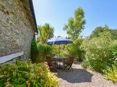 An outdoor patio with a round table and four wooden chairs under a blue umbrella surrounded by plants and a stone wall at Parsonage Farm Cottage in Newton Ferrers