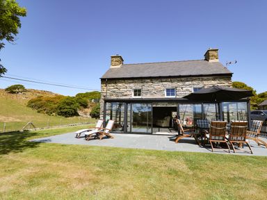 A stone farmhouse with a slate roof and a patio with wooden chairs and a large umbrella at Garth Morthin The Farmhouse in Porthmadog
