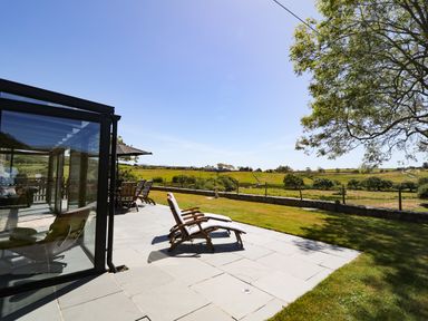 An outdoor patio with lounge chairs and a glass sunroom overlooking a fenced grassy field at Garth Morthin The Farmhouse in Porthmadog