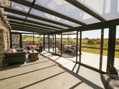 A sunroom with sofas and a wooden coffee table overlooking a patio with a table and chairs at Garth Morthin The Farmhouse in Porthmadog