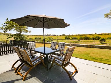An outdoor patio with a table umbrella and six chairs overlooking a grassy field at Garth Morthin The Farmhouse in Porthmadog