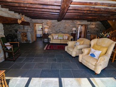 A living room with stone walls wooden beams a sofa two armchairs a dining table and a mounted deer head at Garth Morthin The Farmhouse in Porthmadog