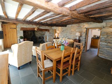 A dining table with chairs and a vase of flowers in a room with exposed wooden beams stone walls and armchairs at Garth Morthin The Farmhouse in Porthmadog