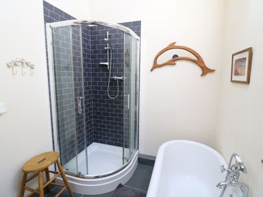 A bathroom with a curved glass shower enclosure tiled with dark subway tiles a white bathtub with chrome fixtures a wooden stool a dolphin-shaped wooden decoration and a framed picture on the wall at Garth Morthin The Farmhouse in Porthmadog