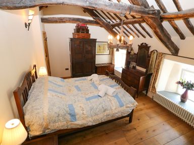 A bedroom with a wooden bed and quilt a wooden wardrobe with suitcases a wooden dresser with mirror and a vase on the window ledge at Garth Morthin The Farmhouse in Porthmadog
