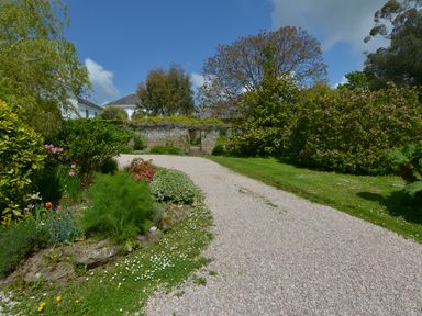 A gravel pathway surrounded by green plants and trees leading to a stone wall with an opening at The Barn in Aveton Gifford