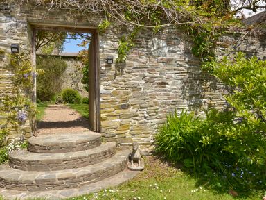 A stone garden wall with an open doorway and steps leading to a gravel path and greenery at The Barn in Aveton Gifford
