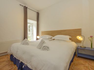 A bedroom with a double bed with white linens and rolled towels near a window with brown curtains and a blue nightstand with a lamp and flowers at The Barn in Aveton Gifford