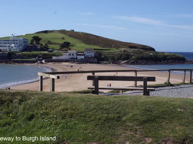 A view of a sandy beach with people walking a wooden bench and green grass in the foreground and buildings and a hill in the background at 3 Tarifa in Bigbury-on-Sea