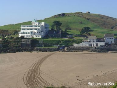 A sandy beach with tire tracks leading to buildings and a green hill in the background at 3 Tarifa in Bigbury-on-Sea