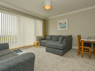 A living room with grey sofas a small wooden table vertical blinds and a dining table with chairs at 3 Tarifa in Bigbury-on-Sea
