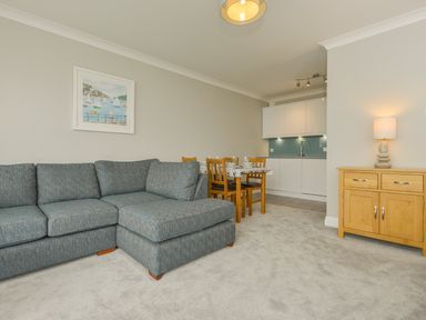 A living room with a grey sofa wooden dining table and chairs and a wooden cabinet with a lamp at 3 Tarifa in Bigbury-on-Sea