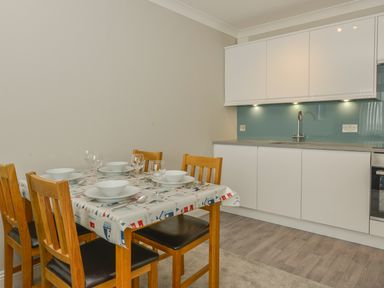A dining table with tableware and wooden chairs next to a kitchen area with white cabinets and a blue backsplash at 3 Tarifa in Bigbury-on-Sea