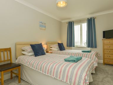 A bedroom with two single beds with striped bedding and dark blue cushions next to a window with blue curtains a wooden chair and a chest of drawers with a television on top at 3 Tarifa in Bigbury-on-Sea