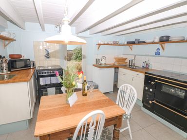 A kitchen with wooden table and two chairs a stove microwave and sink with shelves holding dishes at The Bathing House in Ashprington