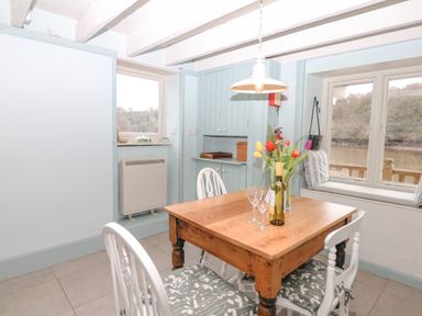 A dining area with a wooden table and four white chairs with cushions by windows showing a water view at The Bathing House in Ashprington
