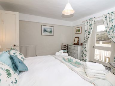 A bedroom with a bed set with pillows and towels near a dresser and chair with a door and floral curtains at The Bathing House in Ashprington