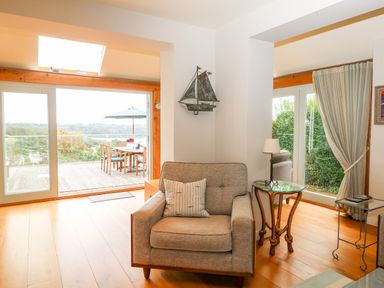 An armchair with a cushion beside a glass table with a lamp in a room with sliding glass doors opening to an outdoor dining area at Dove Cottage in Dittisham
