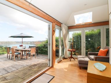 A seating area with a chair ottoman and telescope next to glass doors opening to a wooden deck with a dining table and umbrella at Dove Cottage in Dittisham