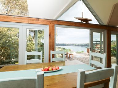 A dining table with fruit bowl and chairs by large glass doors overlooking a deck and water view at Dove Cottage in Dittisham