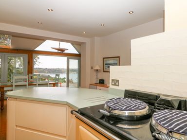 A kitchen with a stove and oven in the foreground and a dining table with chairs near a large window showing a view of a river at Dove Cottage in Dittisham