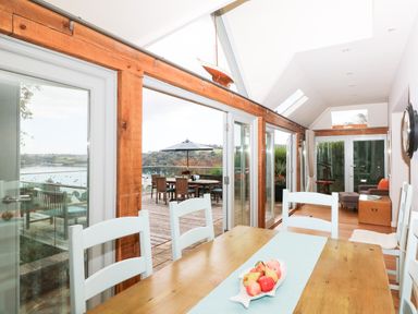 A dining area with a wooden table and white chairs next to glass doors leading to an outdoor deck with a table and umbrella at Dove Cottage in Dittisham