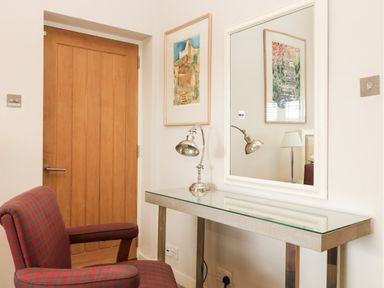 A room with a wooden door a red plaid upholstered chair a glass top desk with a metal lamp and a large mirror on the wall at Dove Cottage in Dittisham