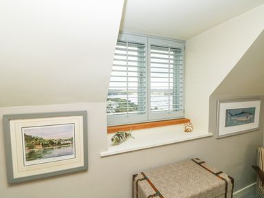 A window with shutters and decorative fish figures on the sill between framed pictures on slanted walls above a patterned storage chest at Dove Cottage in Dittisham
