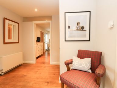 A hallway with wooden floor a plaid armchair with a relax pillow a framed photo on the wall and a dresser with a TV visible through a doorway at Dove Cottage in Dittisham