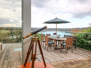 A telescope on a wooden tripod inside looking out to a wooden deck with a dining table and chairs under a large umbrella at Dove Cottage in Dittisham