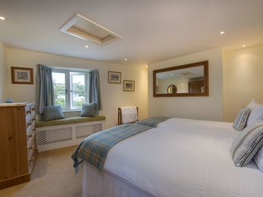 A bedroom with two beds a wooden dresser a window seat with cushions and a large wall mirror at Junket Cottage in Noss Mayo