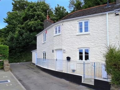 A white stone house with metal railing along the front on a curved road with trees in the background at Junket Cottage in Noss Mayo