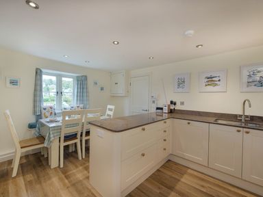 A kitchen and dining area with a table set for four near a window and white cabinets with a sink at Junket Cottage in Noss Mayo