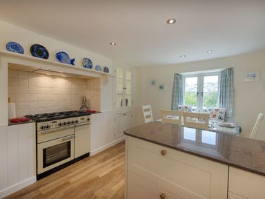 A kitchen with a stove, countertop, dining table set near a window with curtains at Junket Cottage in Noss Mayo