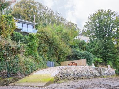 A stone terrace with a wooden picnic table surrounded by dense greenery and a modern house on a hill at Junket Cottage in Noss Mayo