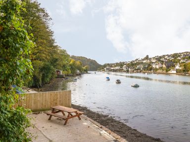 A picnic table on a concrete patio by a river with boats and houses on the opposite bank at Junket Cottage in Noss Mayo