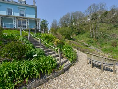 A gravel path with a wooden bench and stairs surrounded by plants leading to a blue house at Woodlands Dartmouth