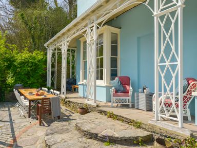 An outdoor patio area with a wooden dining table surrounded by chairs and two cushioned armchairs beside a light blue building with white framed windows at Woodlands in Dartmouth