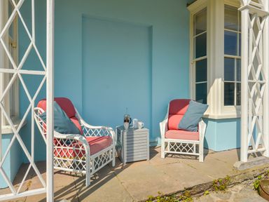 A porch with two white wicker chairs with red cushions and blue pillows and a small gray table with coffee items at Woodlands in Dartmouth