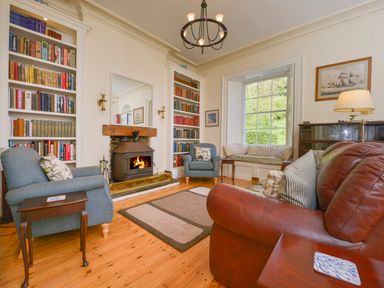 A sitting room with a lit stove fire two armchairs a leather sofa built in bookshelves and a window seat at Woodlands in Dartmouth
