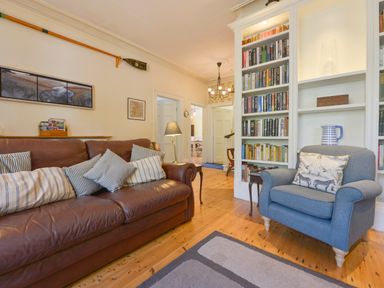 A living room with a brown leather sofa and several striped cushions a blue armchair with a patterned cushion a tall white bookshelf with books a small wooden side table a floor lamp and wooden flooring at Woodlands in Dartmouth