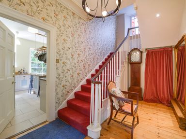 A hallway with red-carpeted stairs floral wallpaper a wooden chair a grandfather clock and a large mirror at Woodlands in Dartmouth