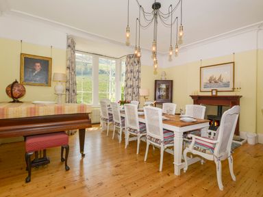 A dining room with a long table and white chairs a grand piano and a fireplace at Woodlands in Dartmouth