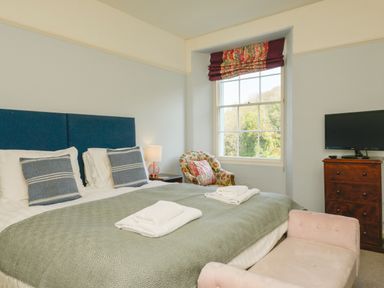 A bedroom with a bed with blue headboard and green blanket a floral armchair a wooden chest of drawers with a TV and a window at Woodlands in Dartmouth